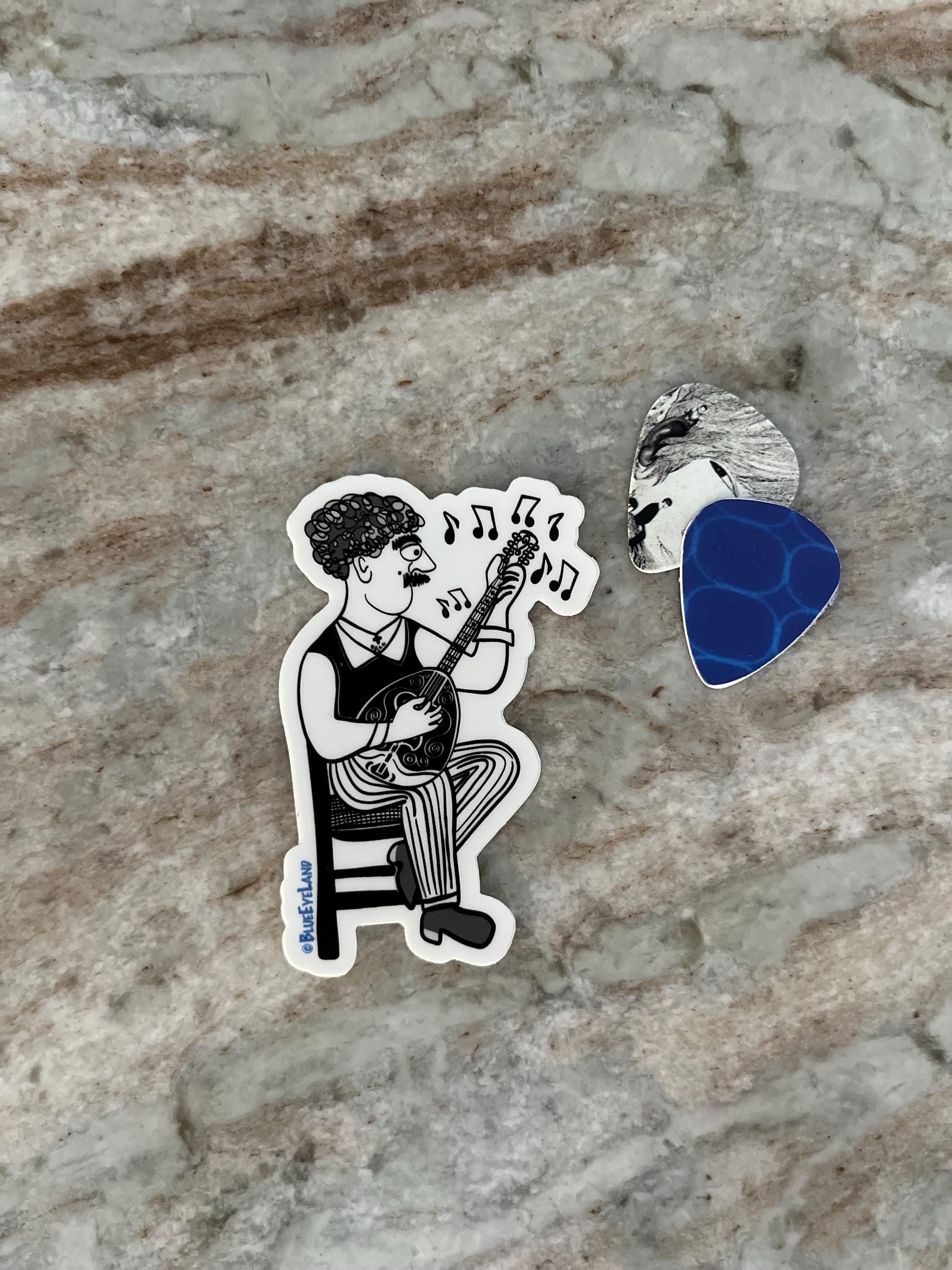 Sticker of a person playing bouzouki with music notes, a guitar pick, and a small silver object on a textured stone surface.