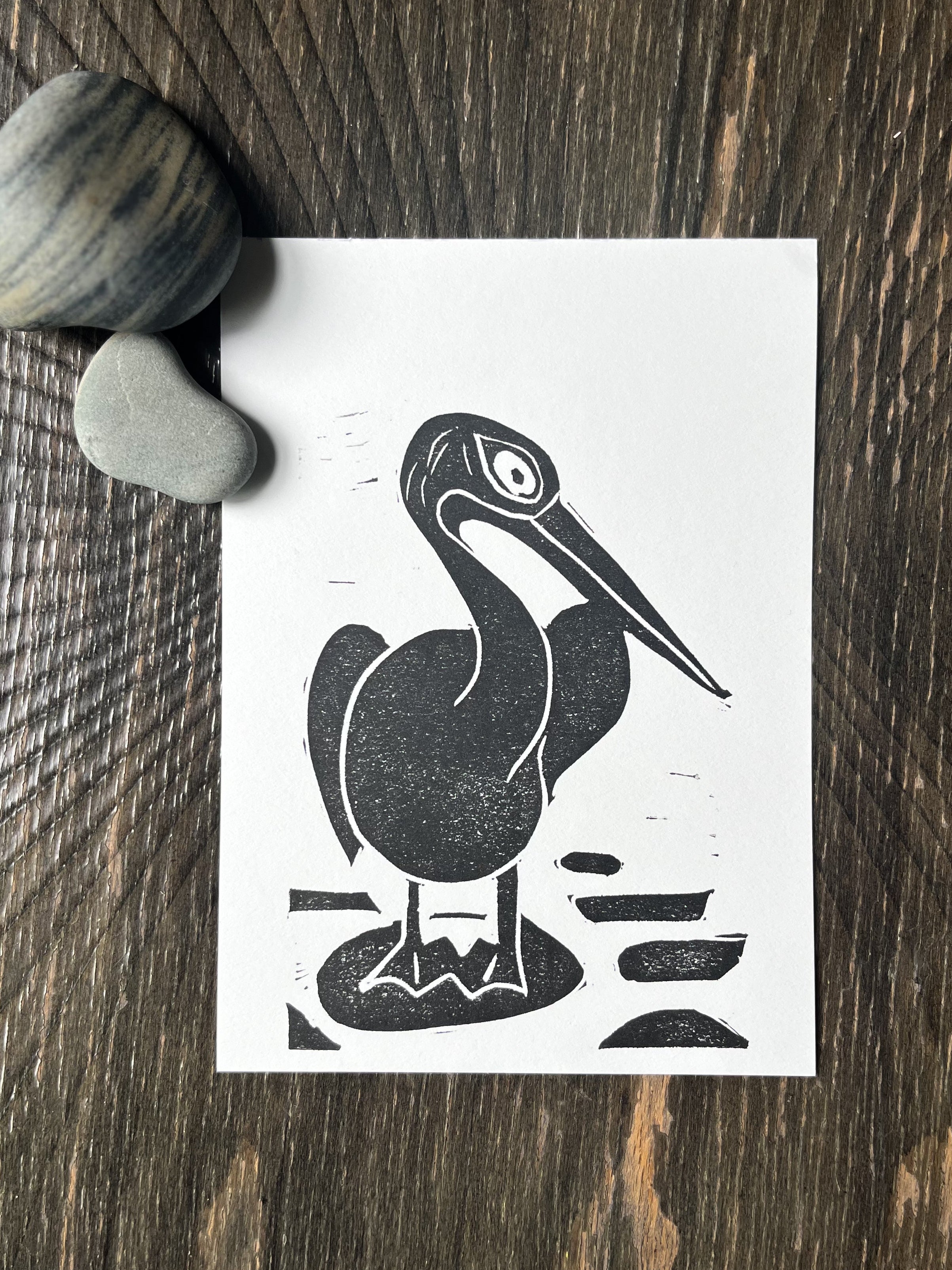 Black and white illustration of a pelican on a white card, placed on a wooden surface with pebbles.